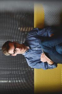 A young man in glasses writes in a notebook while sitting on a stylish couch indoors.